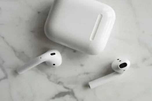 a single premium wireless earbud resting on its open charging case on a wooden desk next to a smartphone, soft morning window light, extreme close-up, editorial product photography, shallow depth of field, no people
